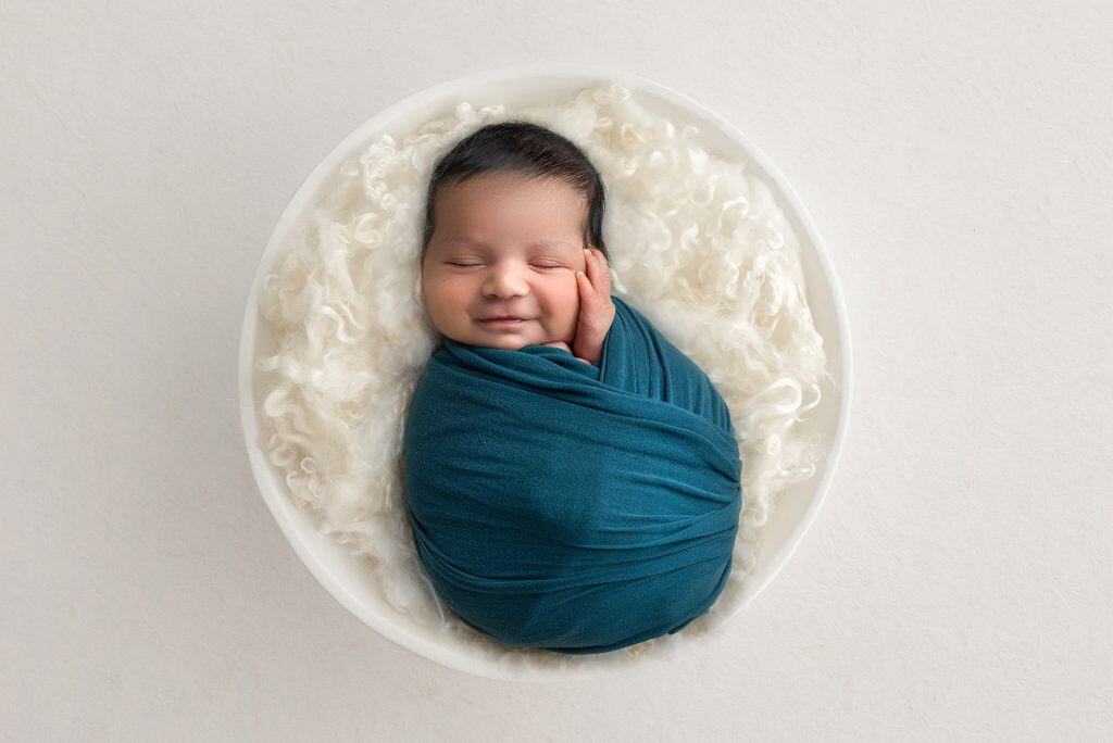 newborn baby boy swaddled in teal asleep on a fluffy blanket inside a white bowl