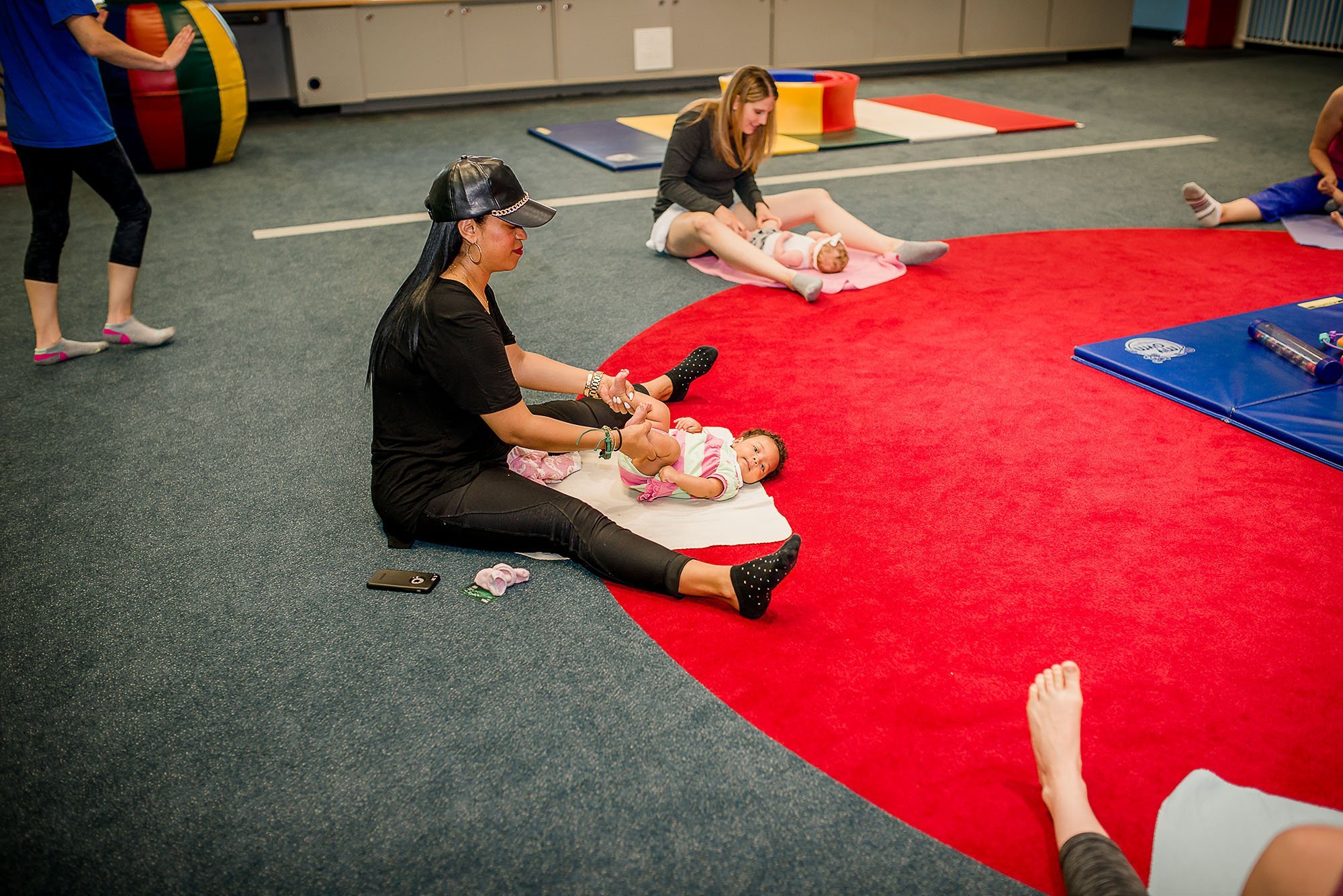 Mom sitting on a carpet at My Gym helping her baby girl bend her knees up and down